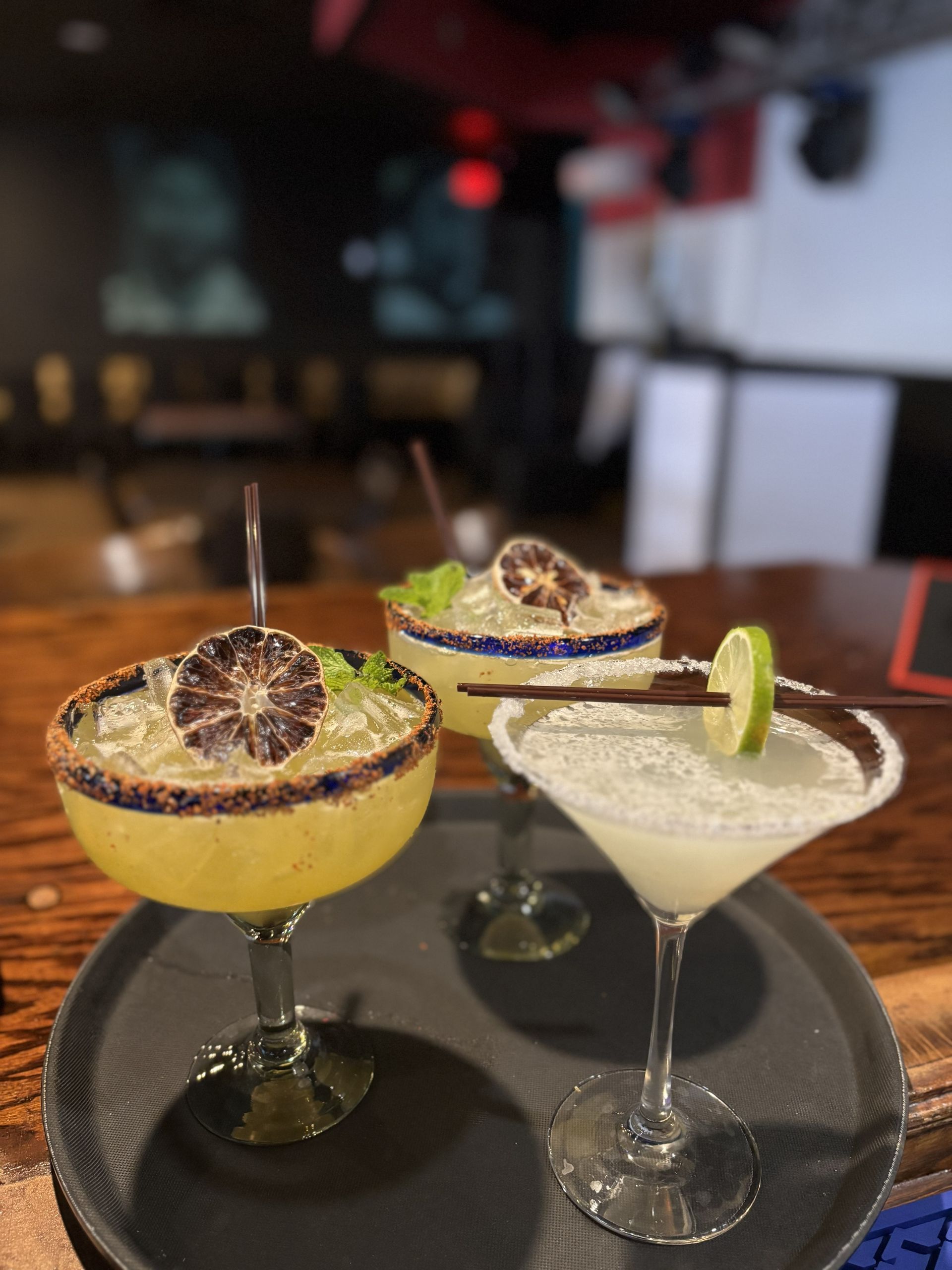 Three margarita cocktails on a tray, various garnishes, salt rims, bar setting.