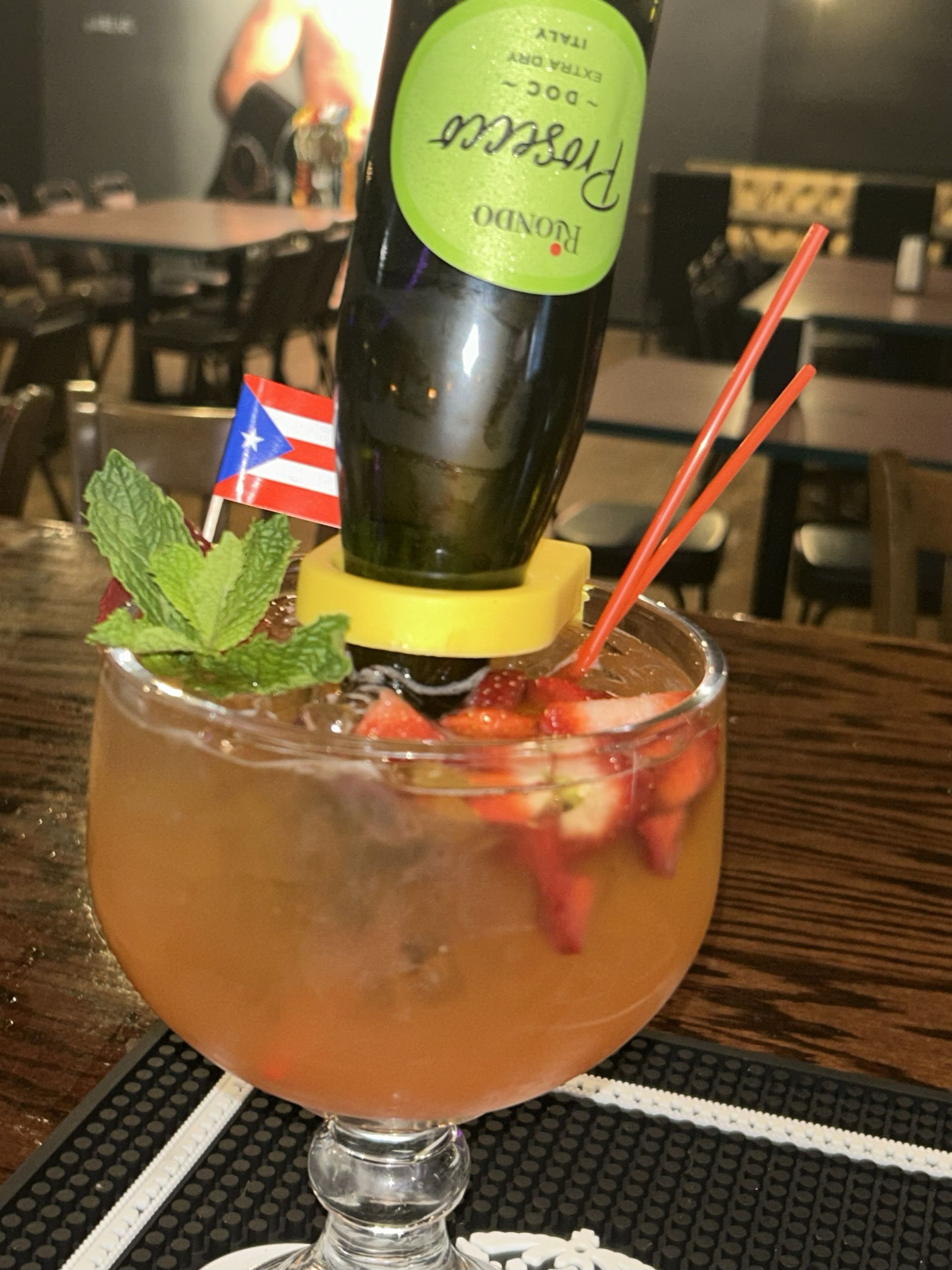 Cocktail in a large glass with a Prosecco bottle upside down, garnished with strawberries, mint, and a Puerto Rican flag.