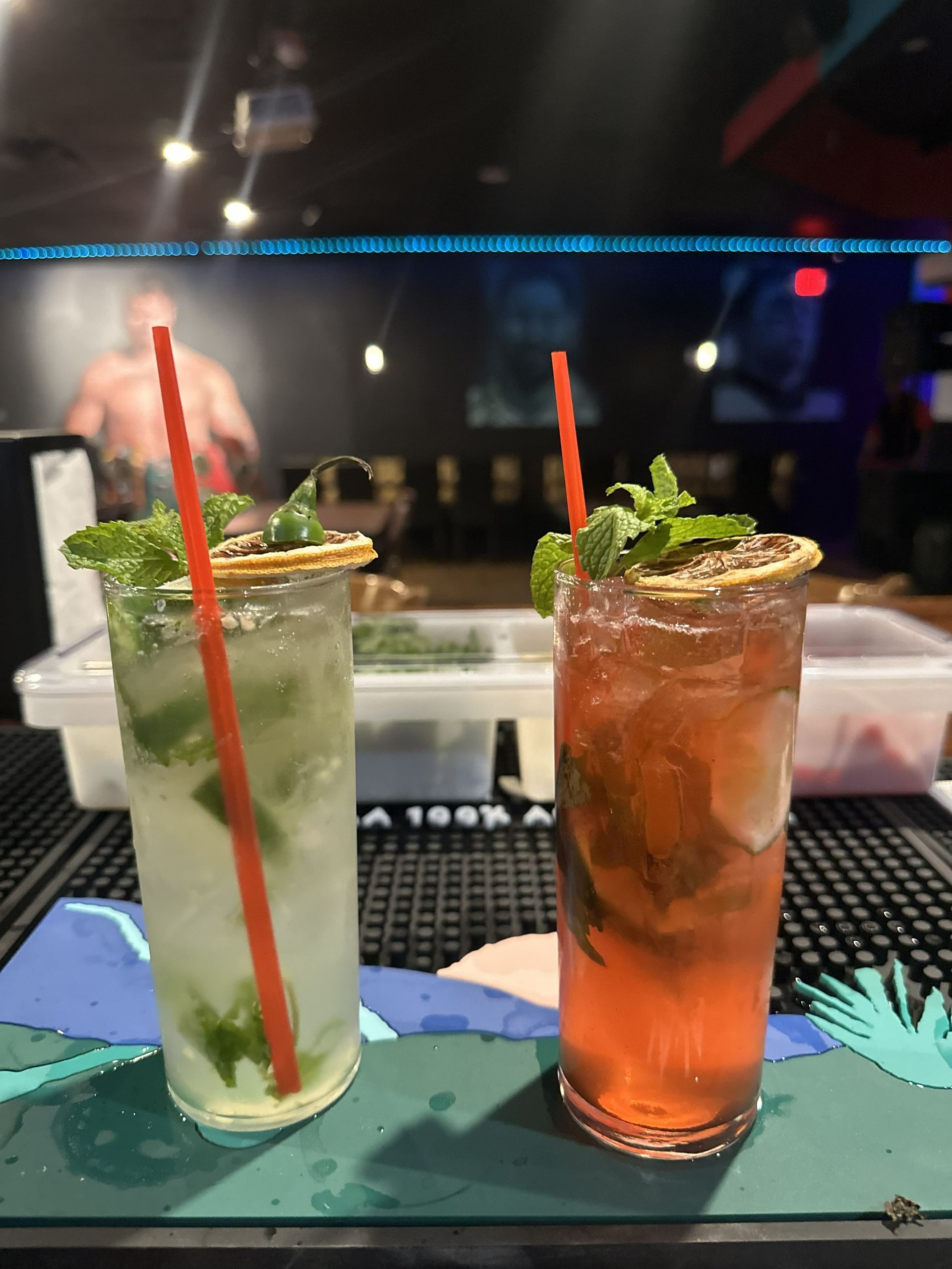 Two cocktails with straws, garnished with mint and citrus on a bar, a person in the background.