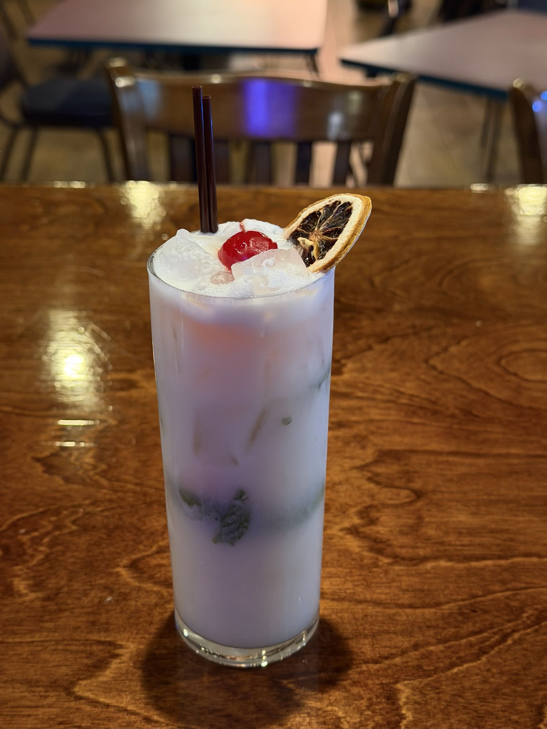 Cocktail in a tall glass, pale purple liquid, topped with whipped cream, cherry, and dried orange slice.
