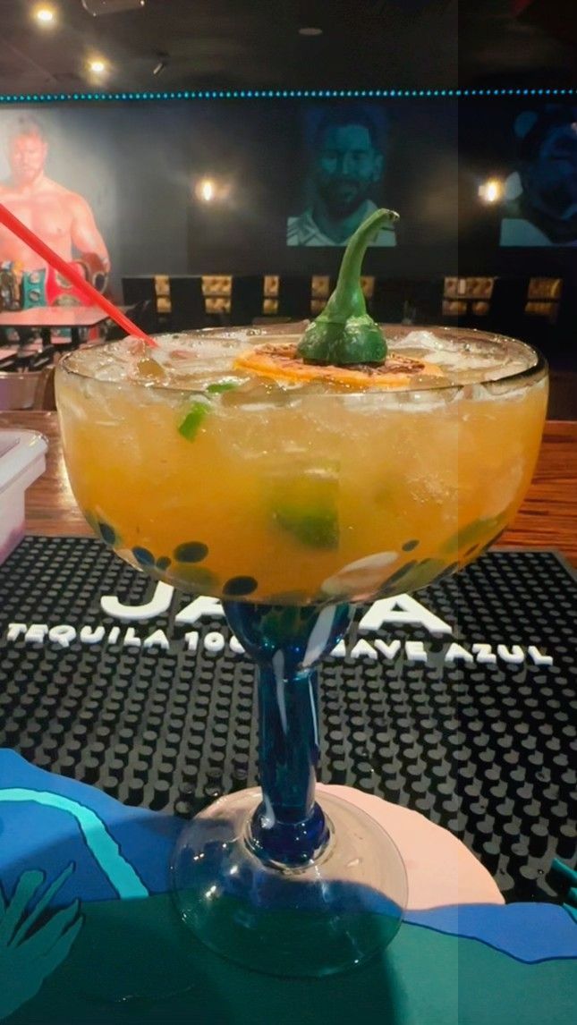Large cocktail in a blue-stemmed glass, garnished with peppers, on a bar with a tequila logo.