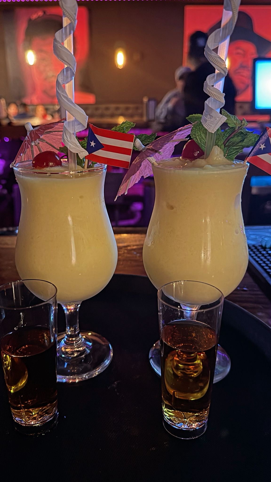 Two piña coladas with cocktail umbrellas, cherry, mint, and Puerto Rican flags, served with shots.