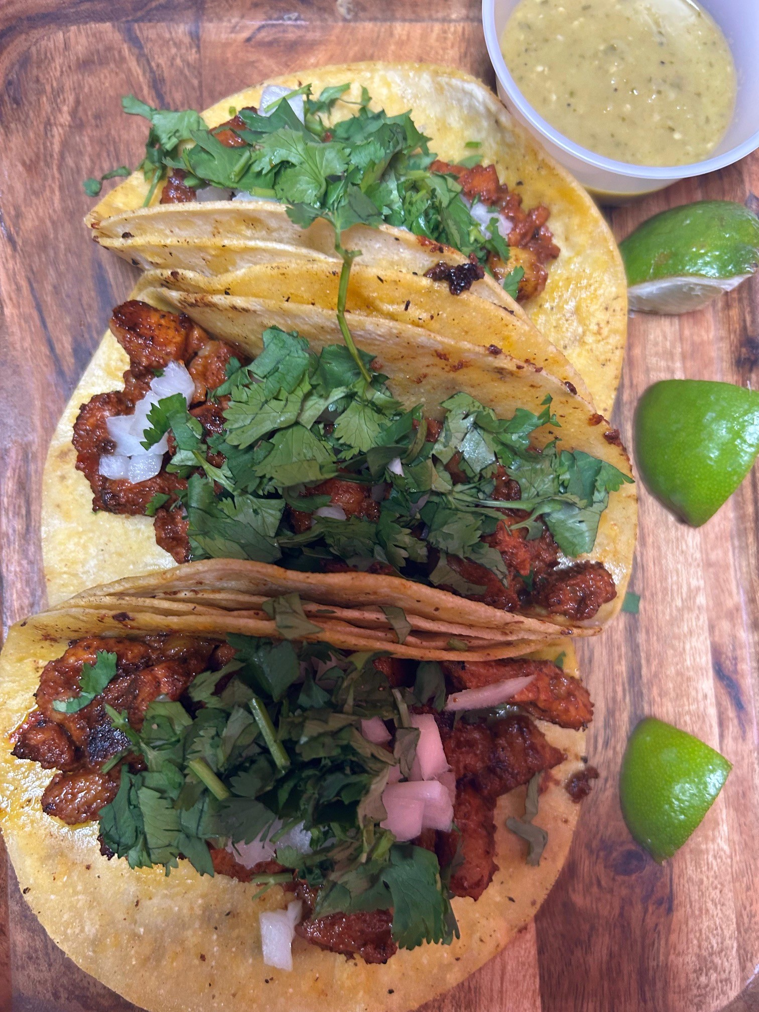 Three tacos on a wooden board, topped with cilantro and onions, with lime wedges and a small bowl of sauce.