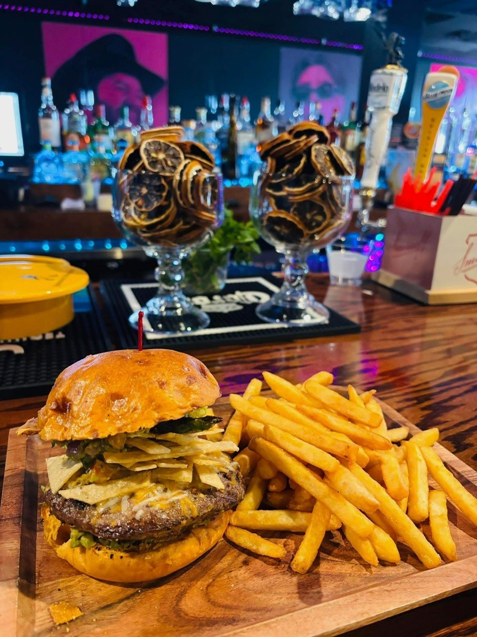 Burger and fries on a wooden board at a bar, next to two glasses with dried citrus.