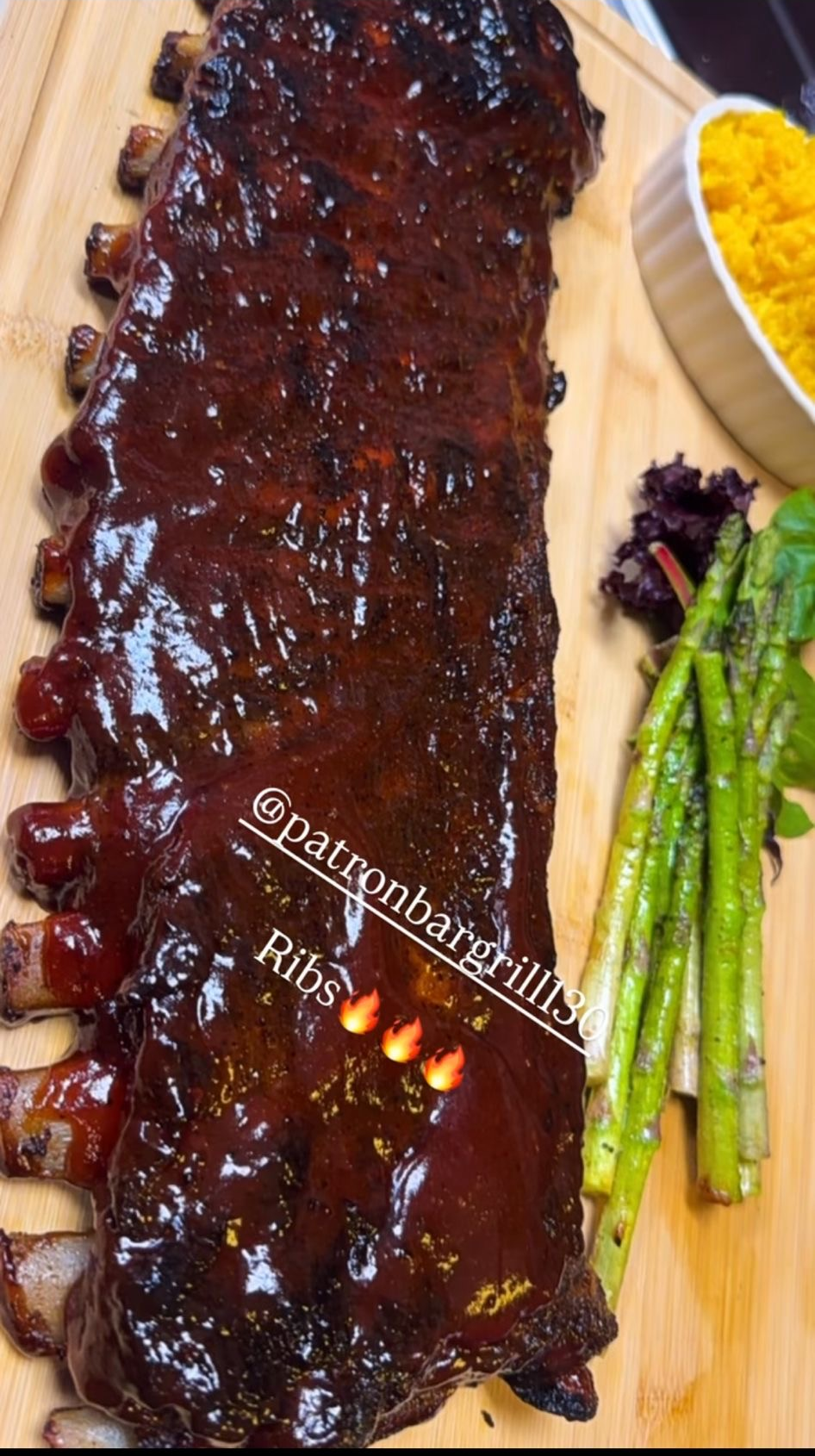 Rack of glazed ribs on a wooden board, with yellow rice and asparagus.