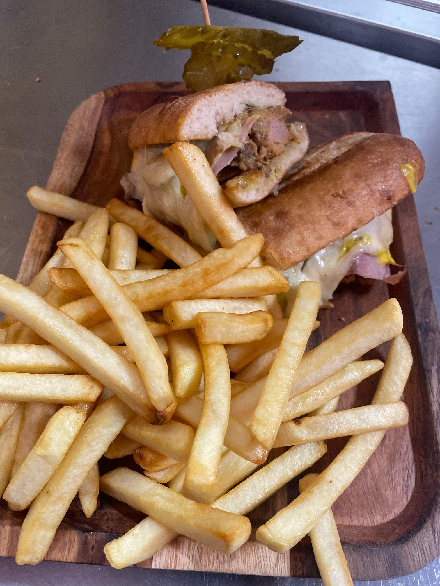 Cuban sandwich with fries on a wooden tray, topped with a pickle.