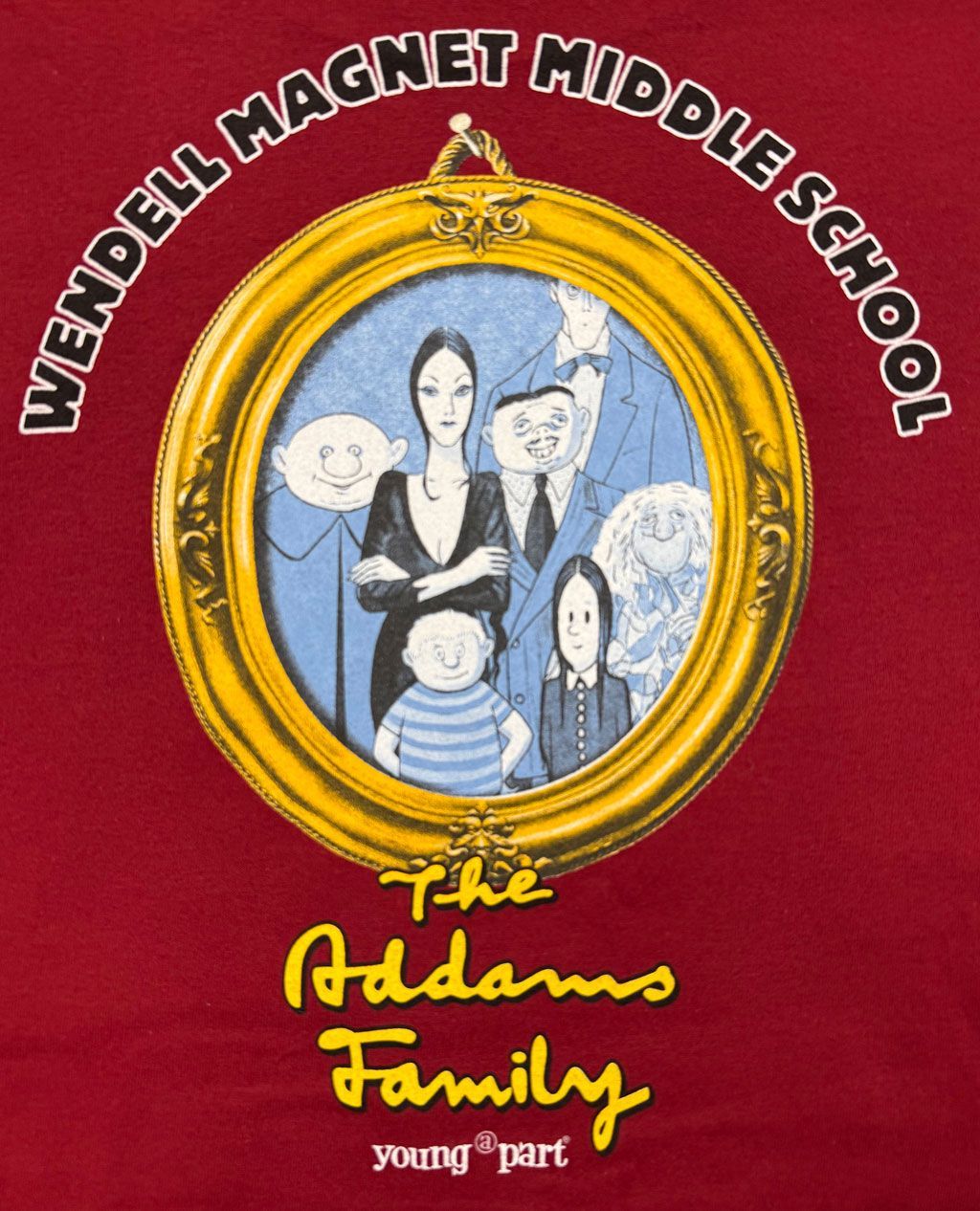 A red T-shirt featuring an Addams Family illustration in an oval gold frame with the text 