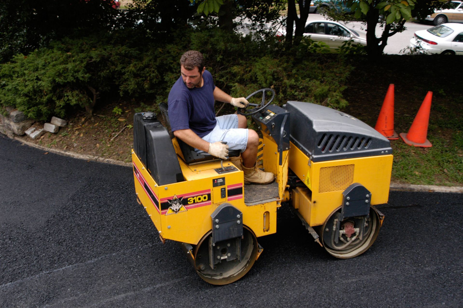 Man riding a small yellow utility cart on a paved road near orange cones and greenery