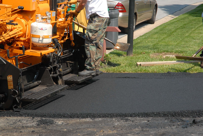 Road paving machine laying fresh black asphalt beside a curb and parked car
