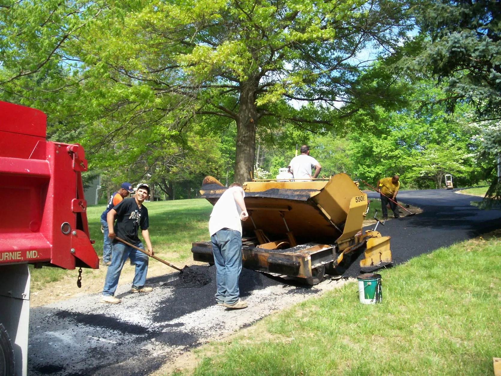 Road repair crew paving a driveway beside a red truck and yellow paver on a tree-lined street