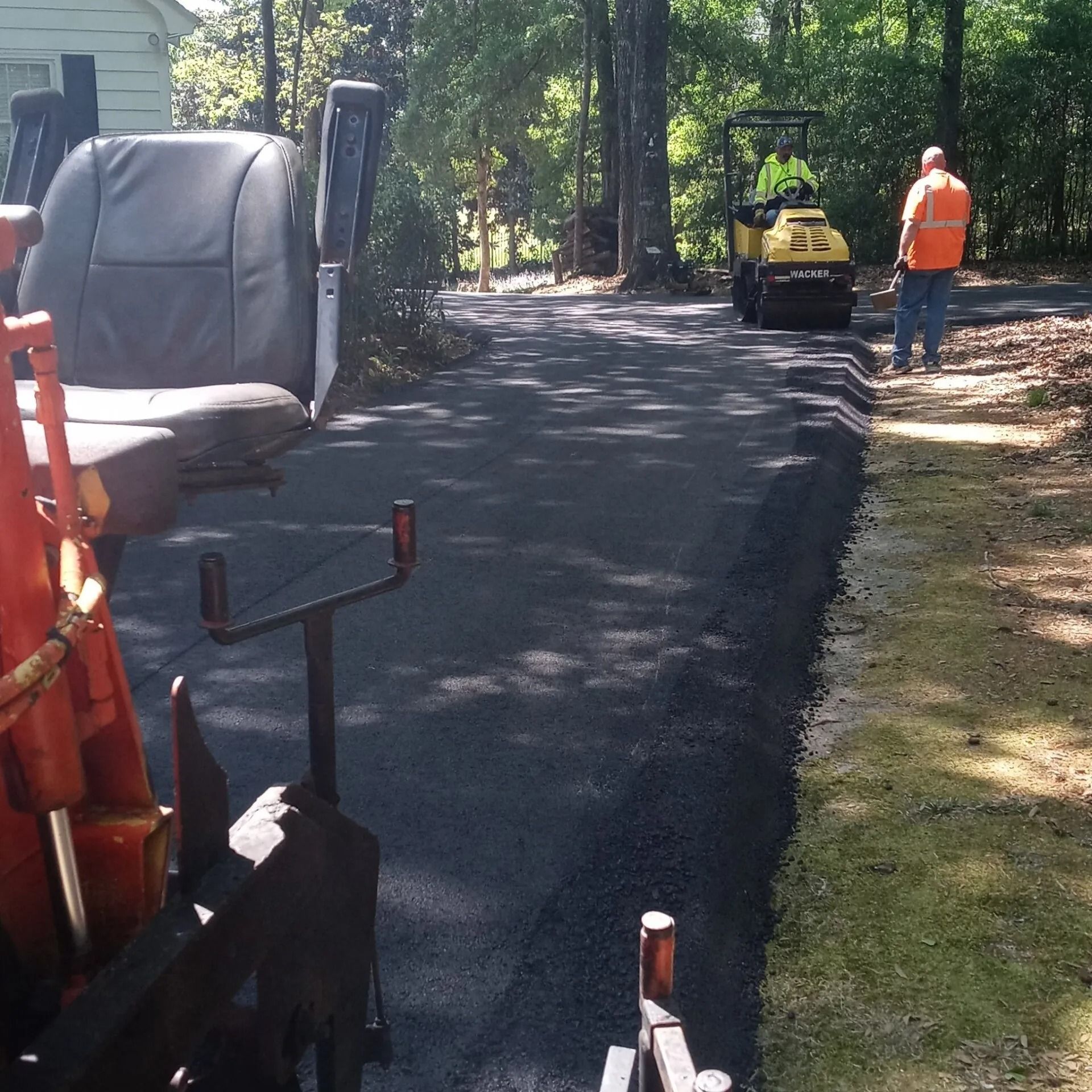 Fresh asphalt driveway being paved, with a worker and compact roller on the right side.
