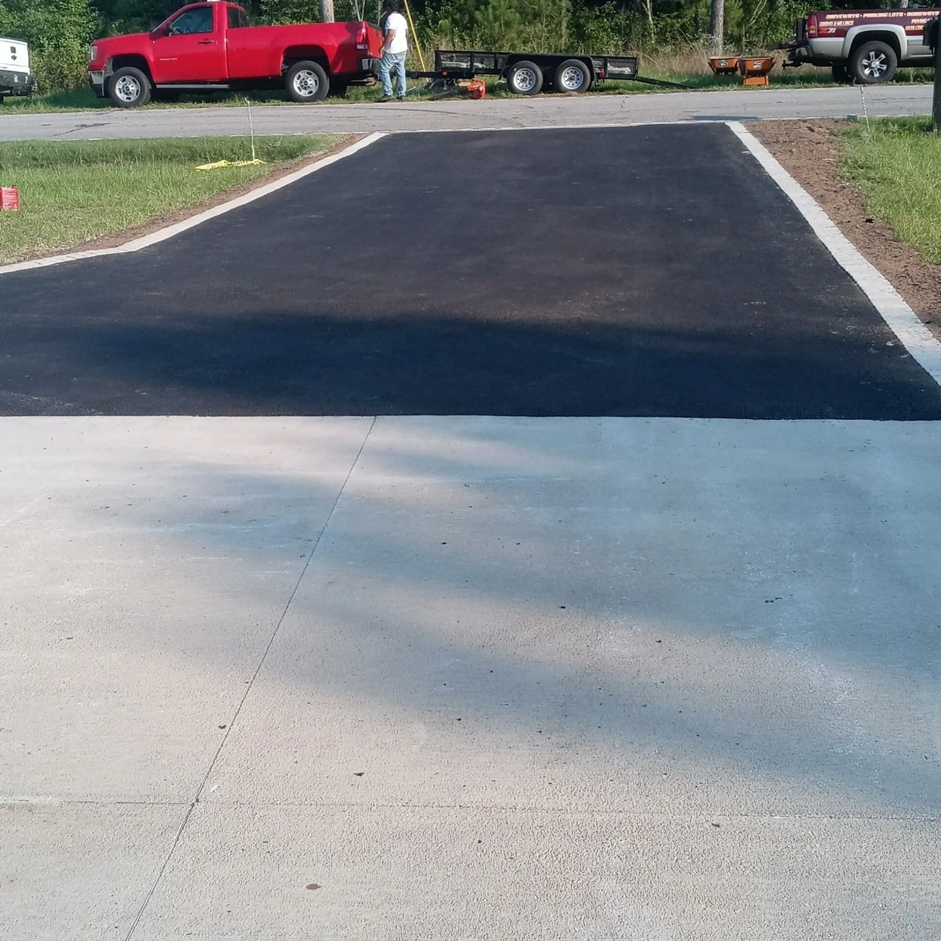 Freshly paved driveway with a dark asphalt section meeting light concrete, with parked trucks in the background