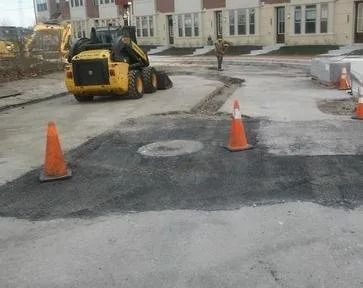 Construction equipment patching a roadway, with orange cones and a worker near buildings.