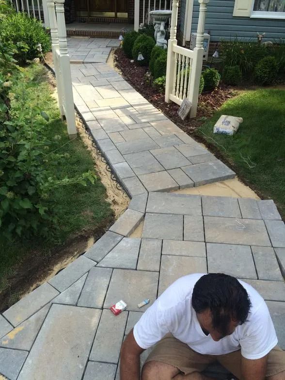 Person kneeling on a stone walkway leading to a house entrance with white railings and greenery nearby