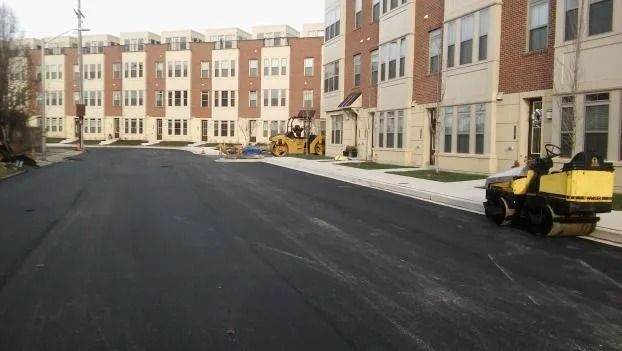 Freshly paved street in front of apartment buildings, with construction equipment parked along the curb