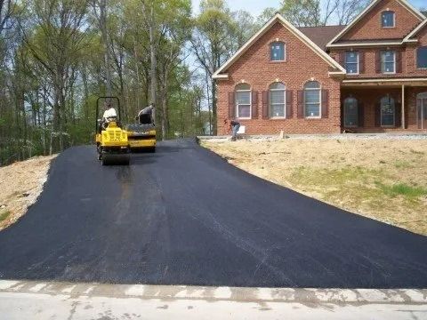 Yellow construction roller paving a new black asphalt driveway beside a brick house