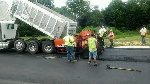 Workers repair a damaged road beside a tipped dump truck with spilled debris.