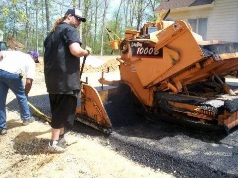 Workers operating a paving machine on a driveway, spreading fresh asphalt beside a house.