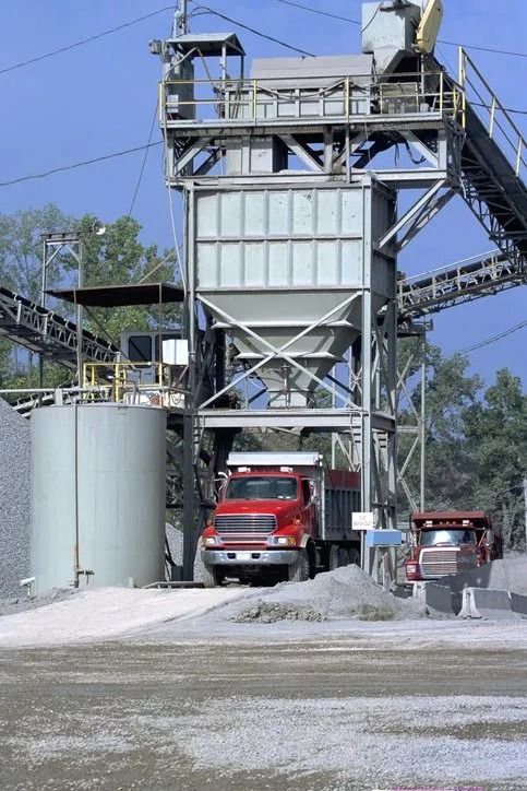Red truck under a concrete batching plant with silos, conveyor, and gravel yard