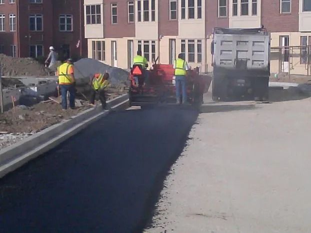 Road workers in safety vests paving a fresh black asphalt street beside a building.