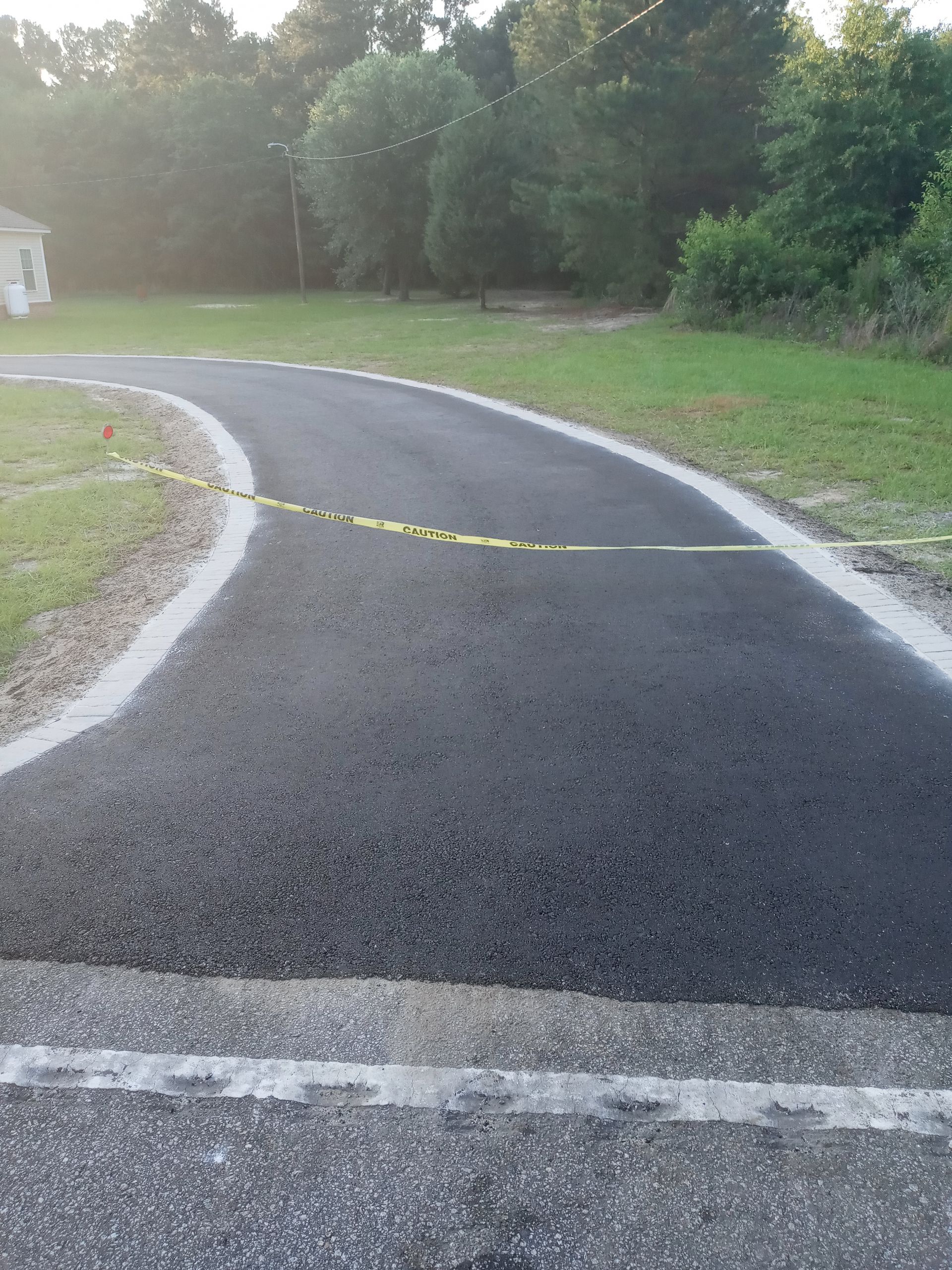 Curved asphalt path in a park, with yellow caution tape across it and trees in the background.