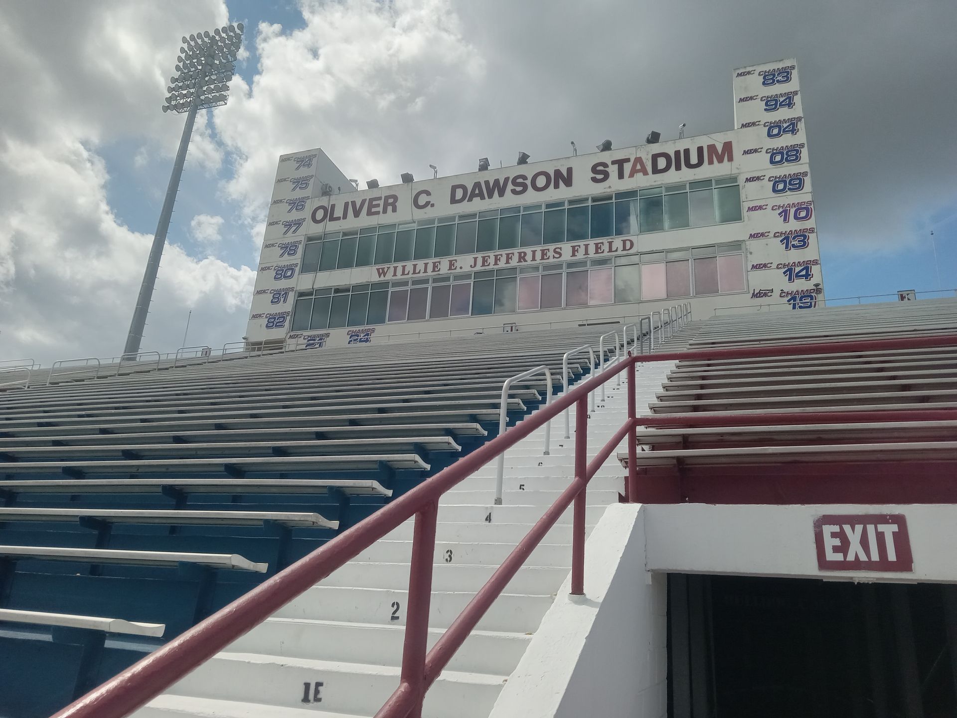 Empty stadium seating leading to Oliver C. Dawson Stadium scoreboard and exit sign under cloudy sky