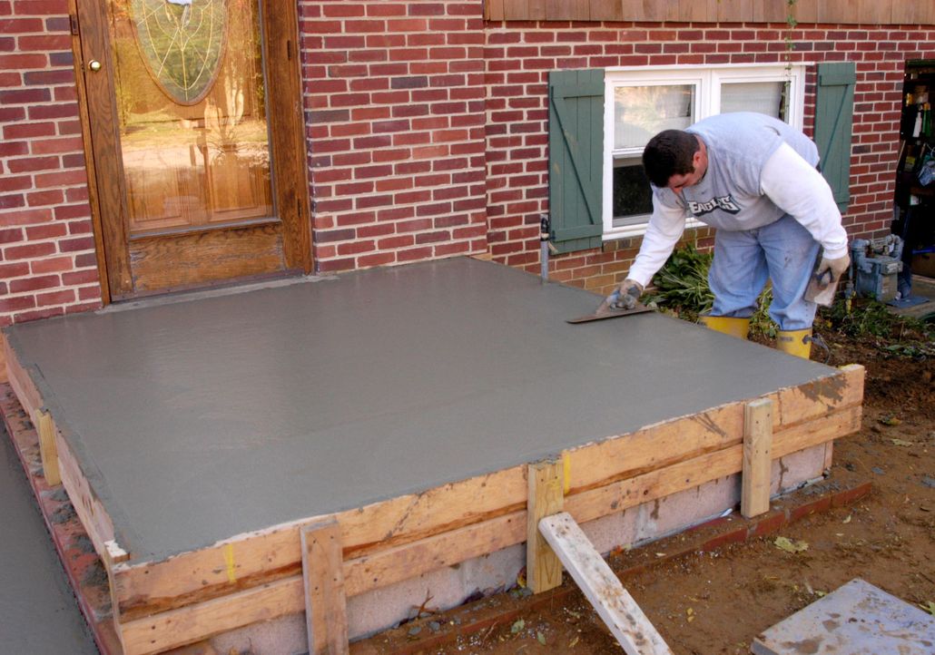 Worker smoothing fresh concrete slab beside a brick house foundation
