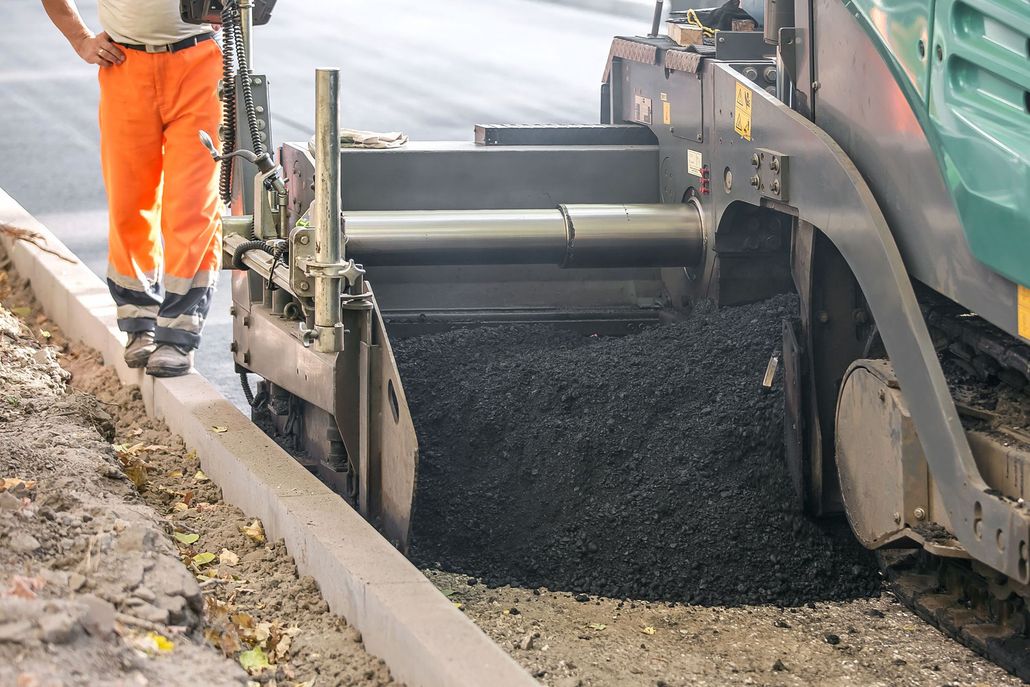 Road paving machine laying asphalt beside a curb, with a worker in orange pants nearby
