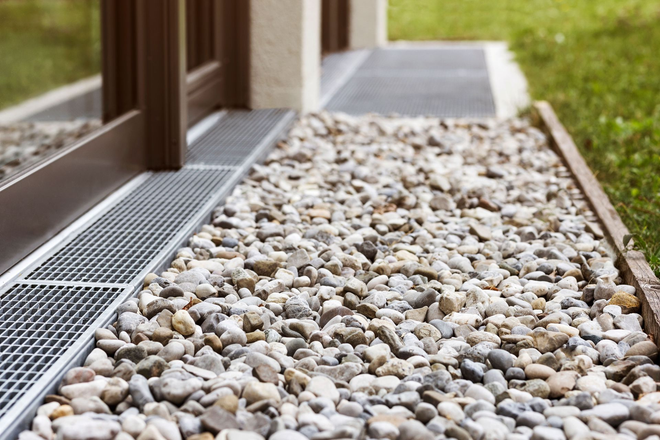 Gravel bed beside a house with a narrow metal drainage channel along the edge