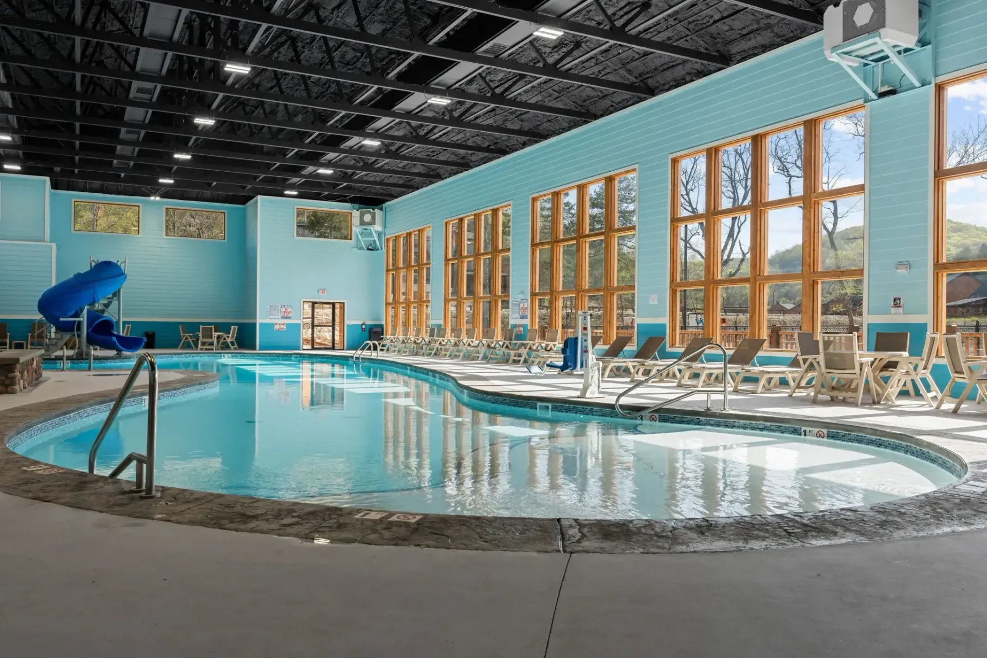 Indoor pool with lounge chairs, large windows, and a blue slide under a high ceiling.