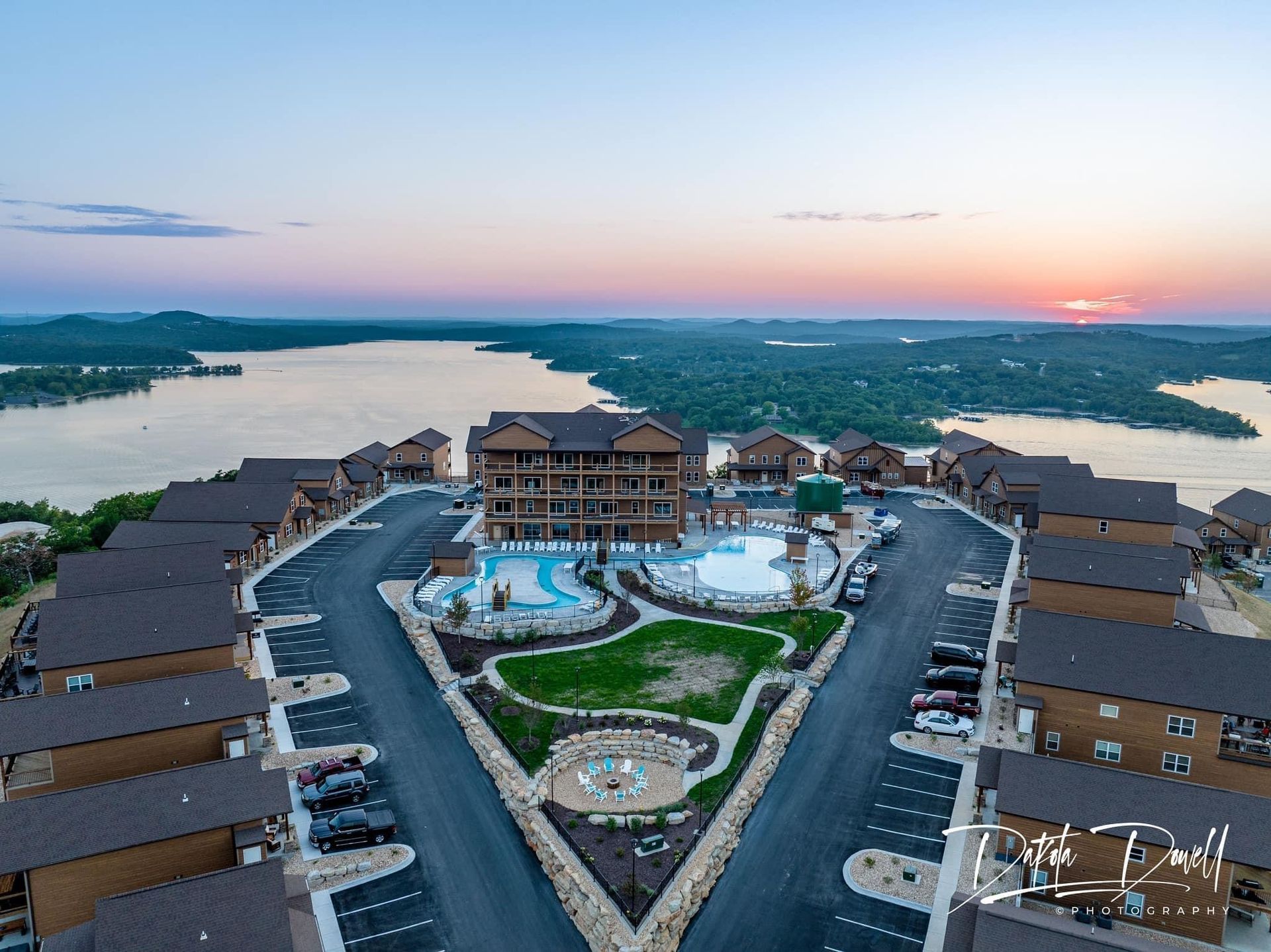 Aerial view of a lakeside resort with a pool, roads, and sunset over the water.
