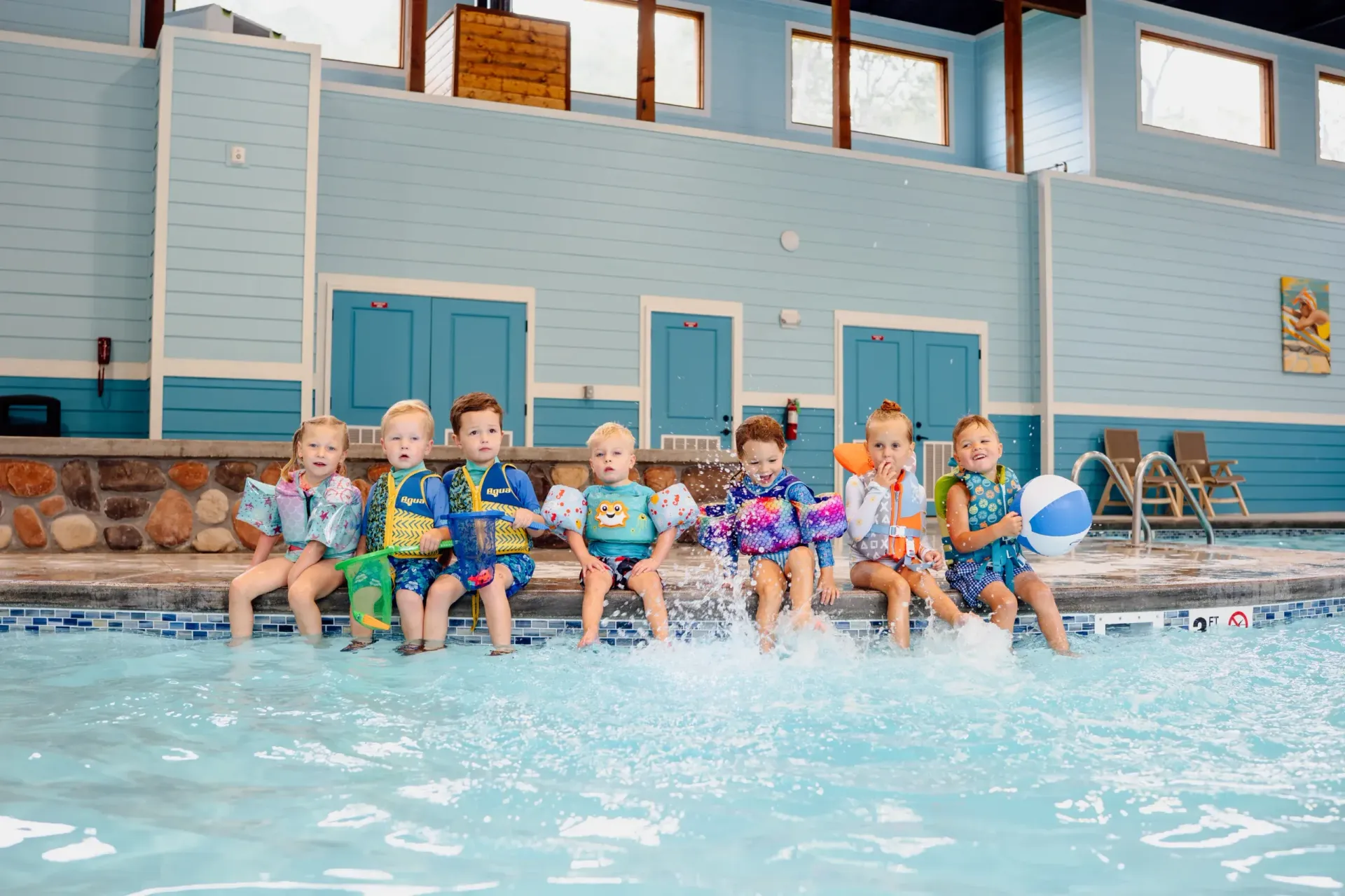 Children sitting by an indoor pool with blue water and light blue walls, holding pool toys and kicking their feet