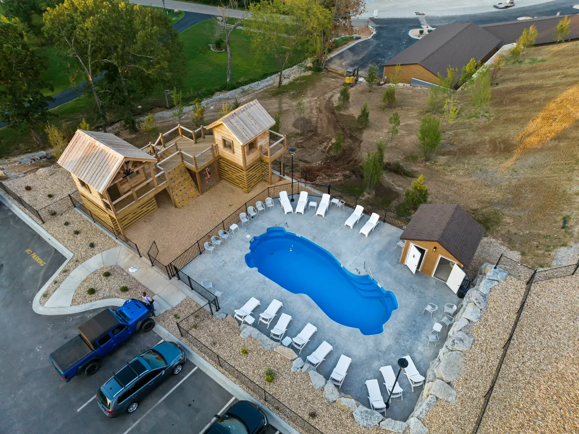 Aerial view of a backyard pool with lounge chairs, small cabanas, and parked cars nearby.