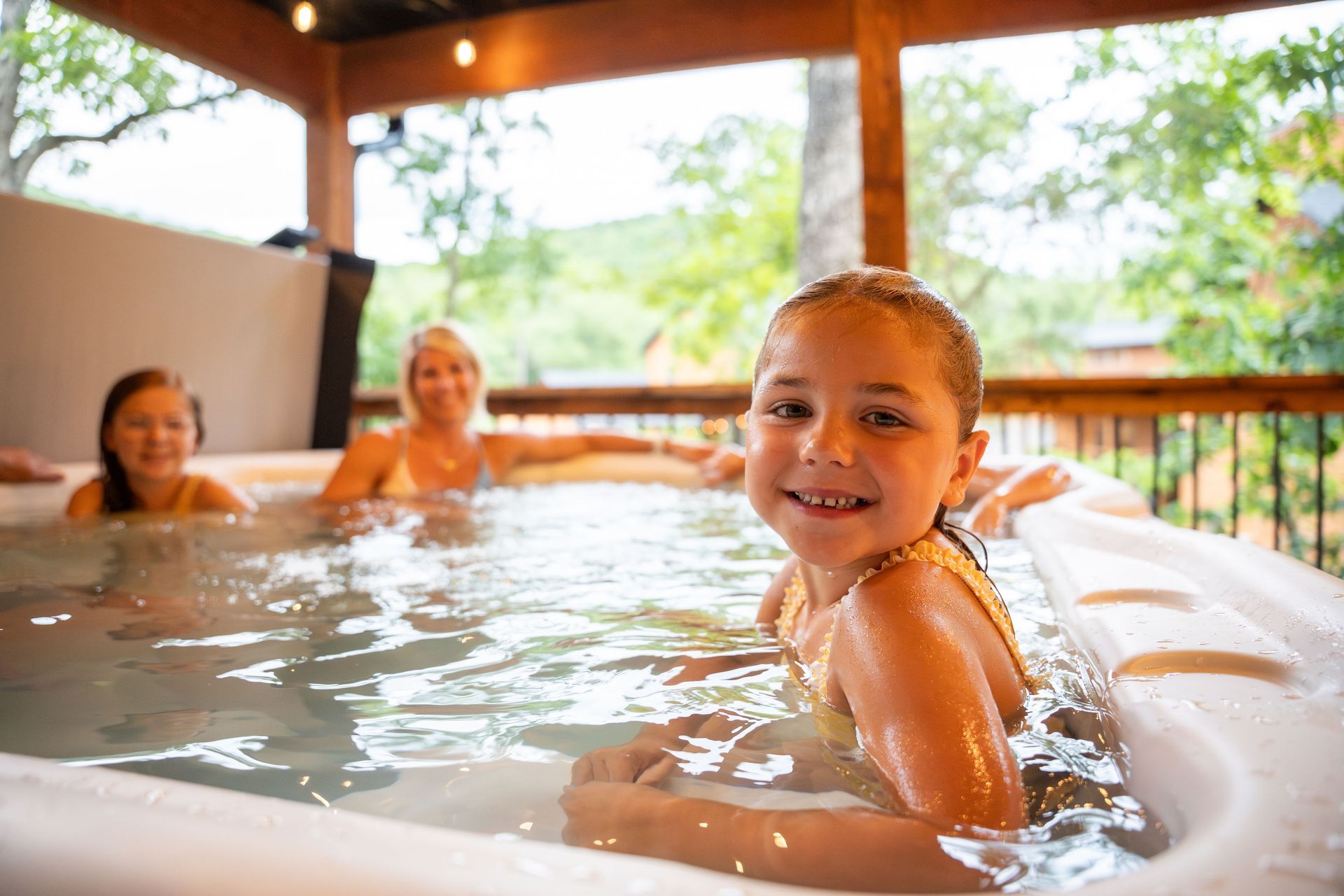 People relaxing in an outdoor hot tub, with a child smiling in the foreground