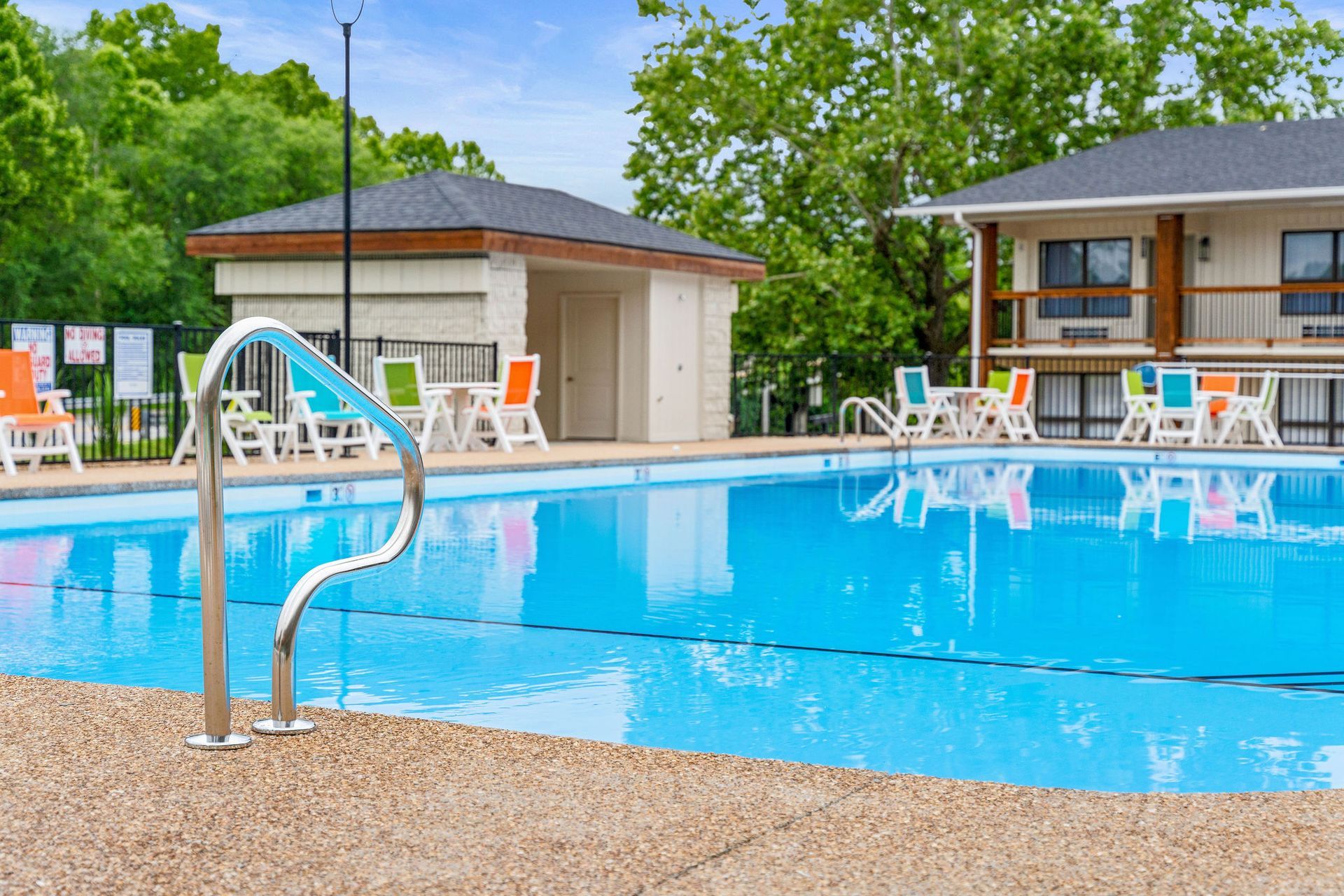 Outdoor swimming pool with blue water, white lounge chairs, and beige pool houses under green trees