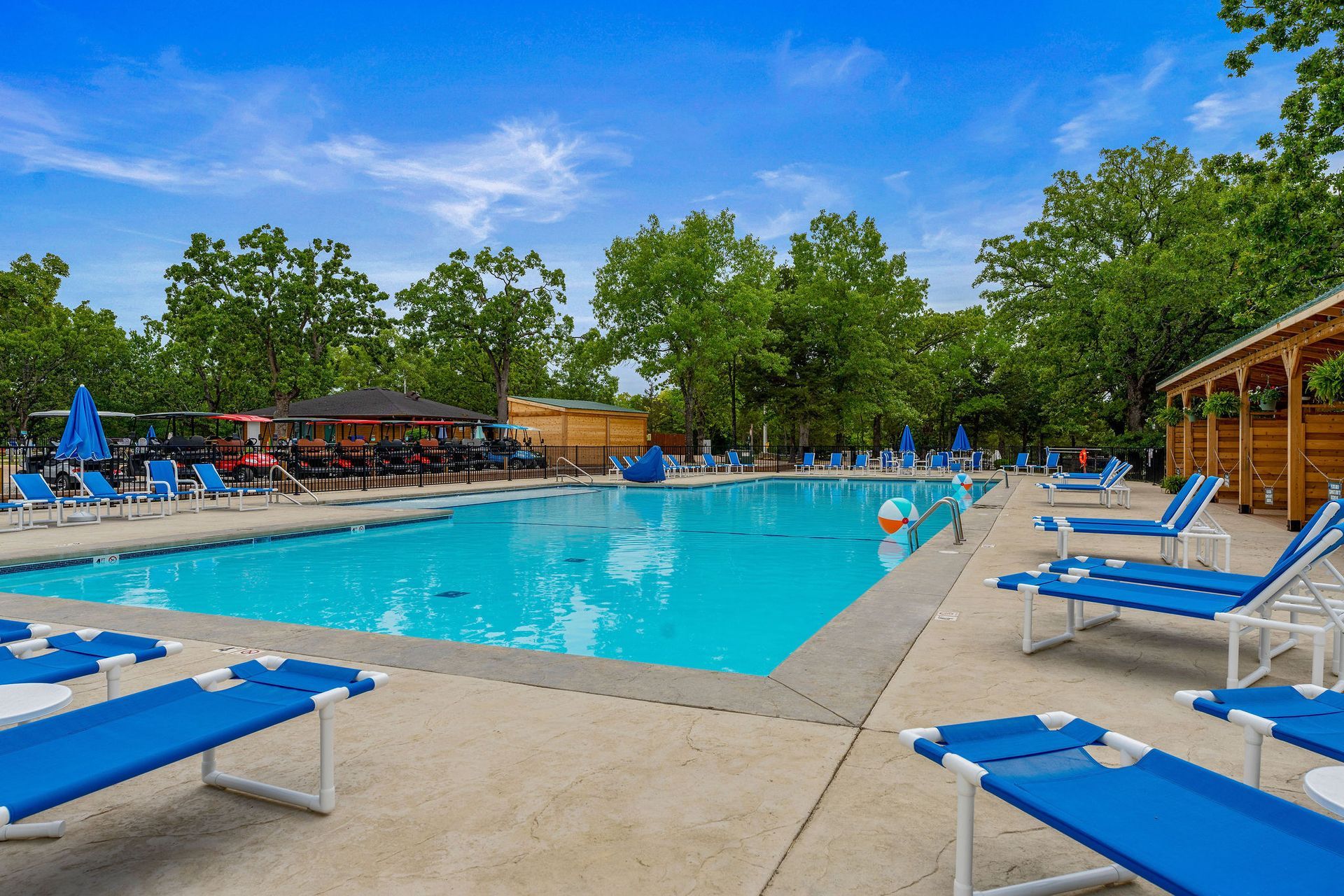 Outdoor swimming pool with blue lounge chairs, umbrellas, and trees under a bright blue sky
