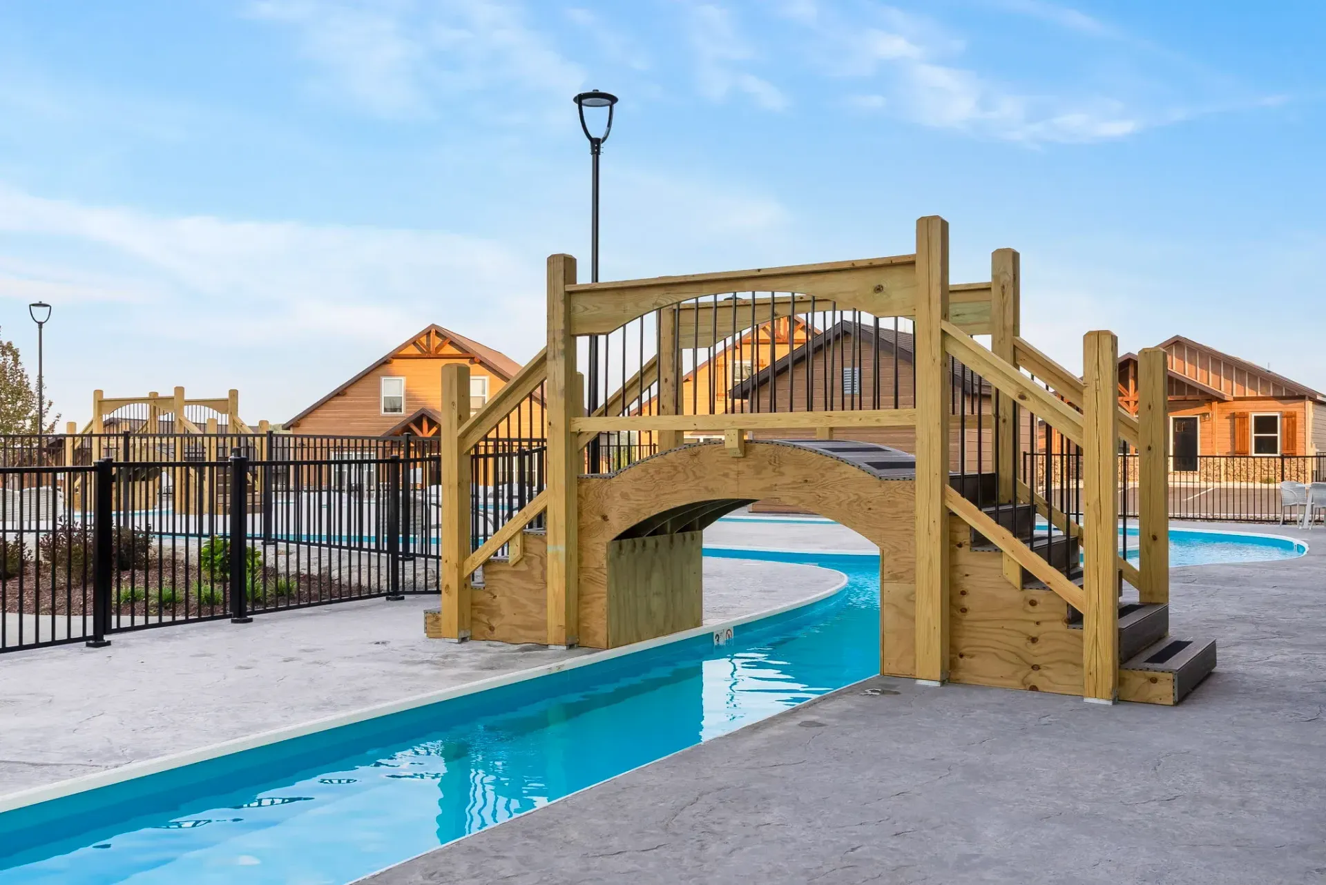 Wooden footbridge over a narrow pool in a residential courtyard, with houses and a black fence nearby