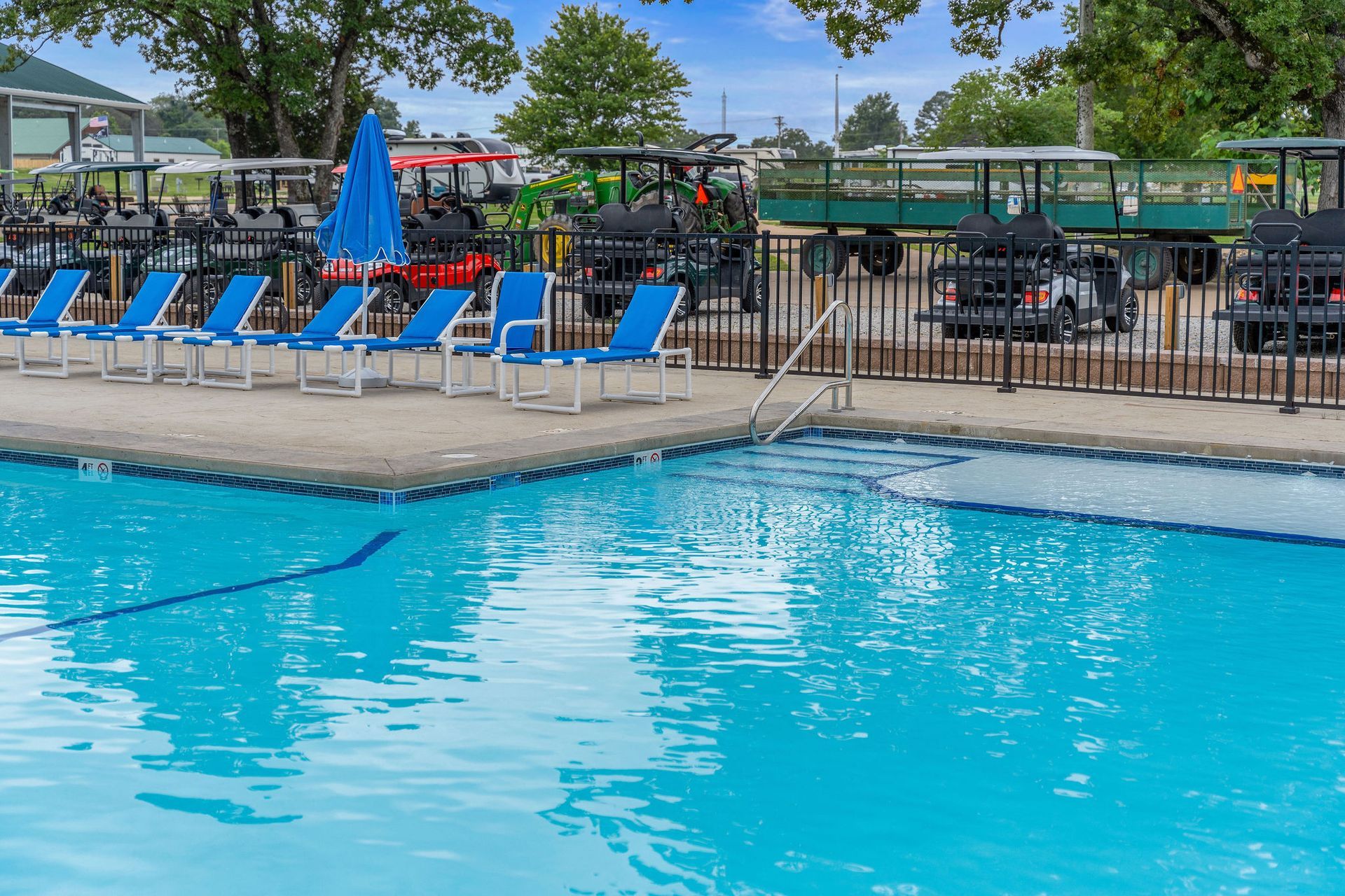 Outdoor swimming pool with blue lounge chairs, umbrellas, and fenced seating area in the background