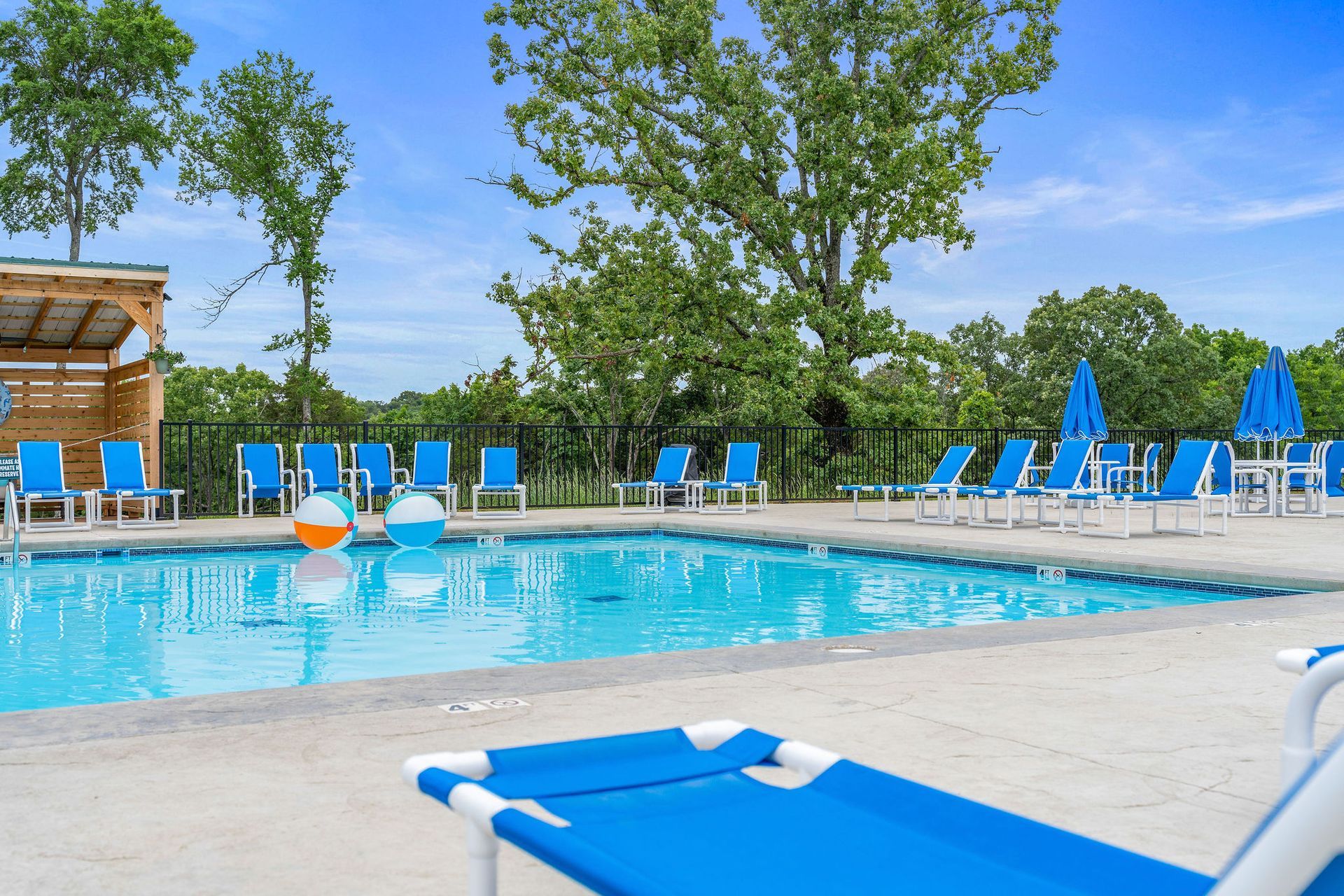 Outdoor swimming pool with blue lounge chairs, clear water, and trees under a sunny sky