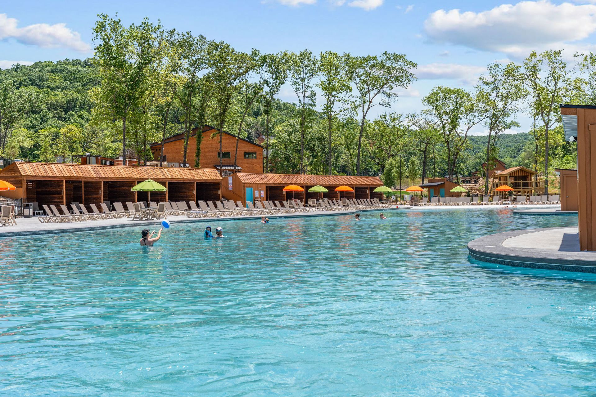 Resort pool with swimmers, orange lounge chairs, and trees under a blue sky