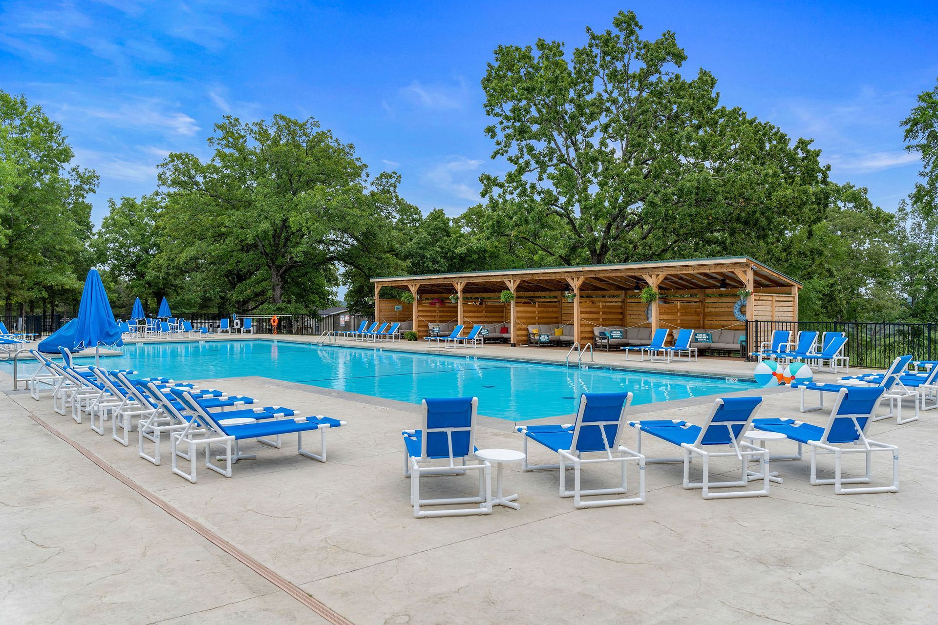 Outdoor pool with blue lounge chairs, umbrellas, and a wooden cabana surrounded by trees