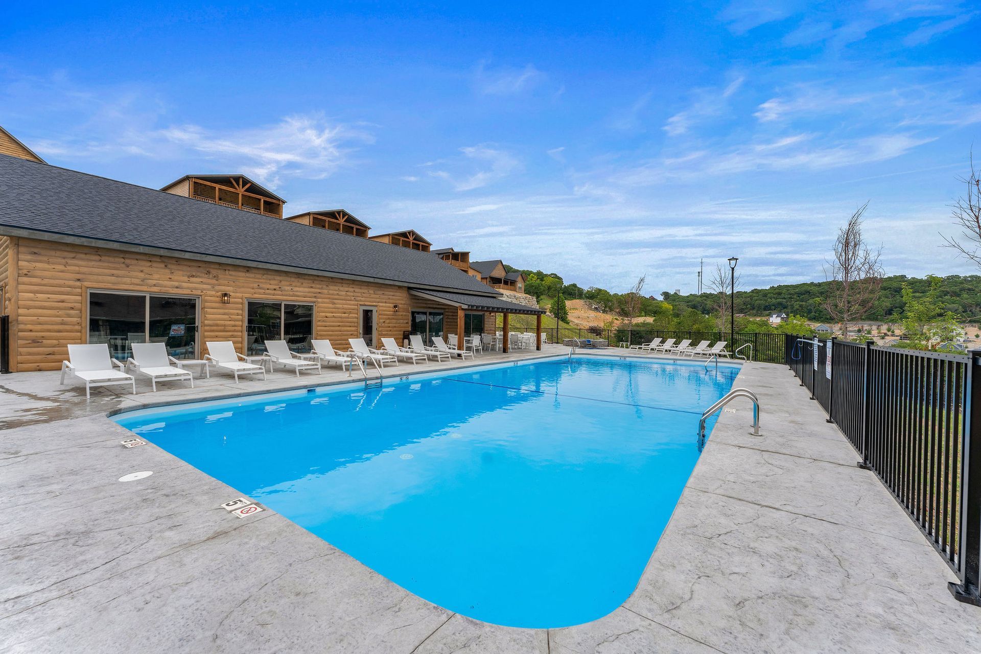 Outdoor swimming pool beside a brick building, with lounge chairs and a black metal fence under a blue sky