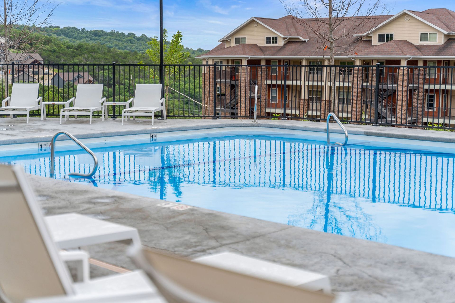 Outdoor swimming pool with white lounge chairs and a wooden fence, with houses and hills in the background