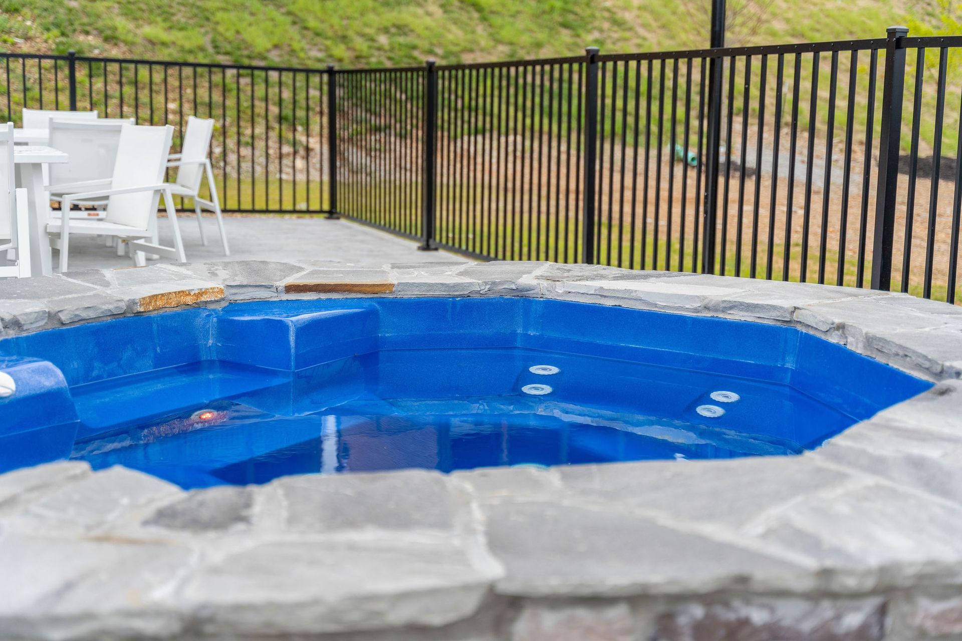 Small backyard pool with blue water, stone border, white chairs, and a black metal fence