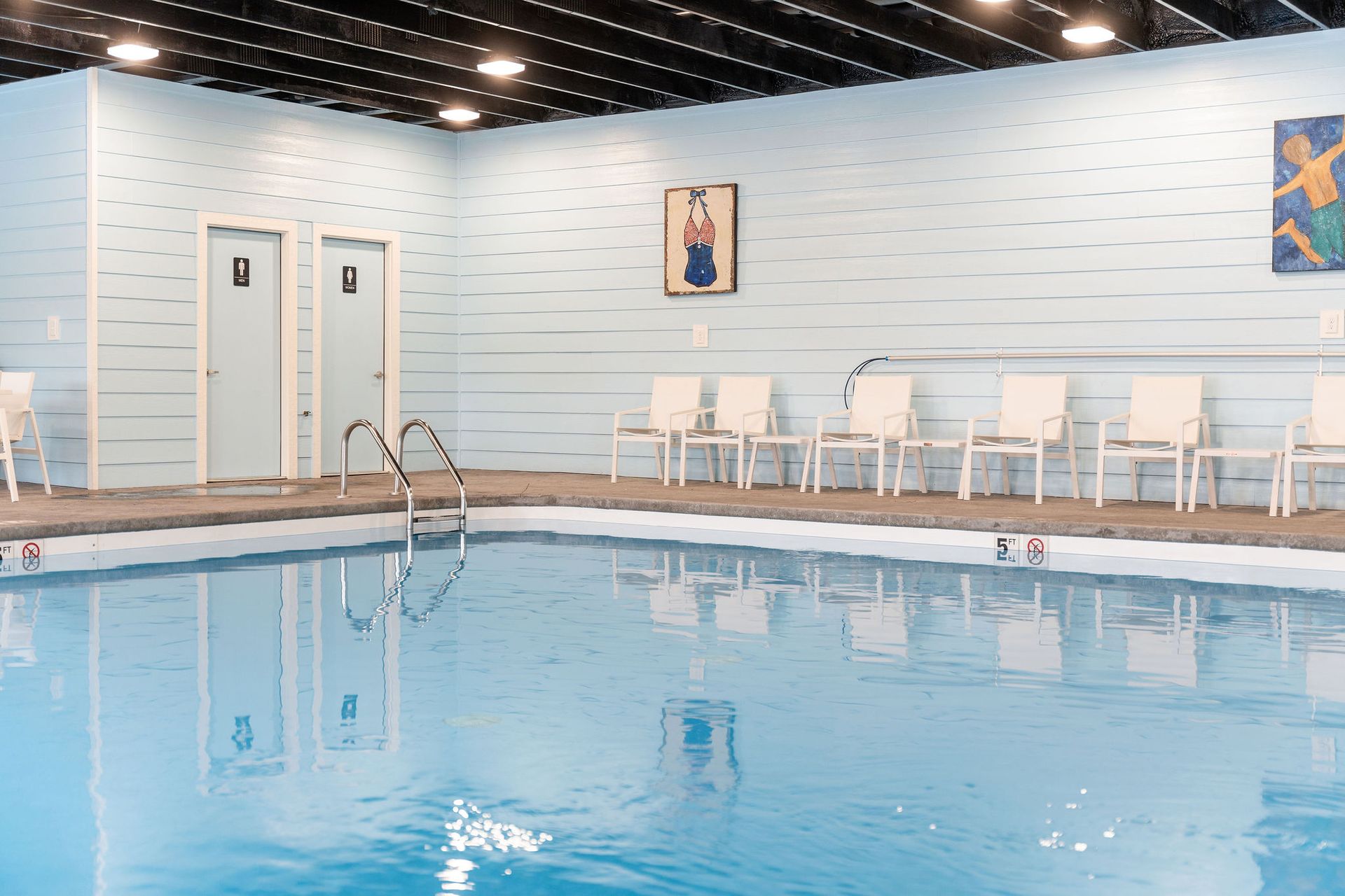 Indoor swimming pool with blue water, white lounge chairs, and ladders along a tiled wall