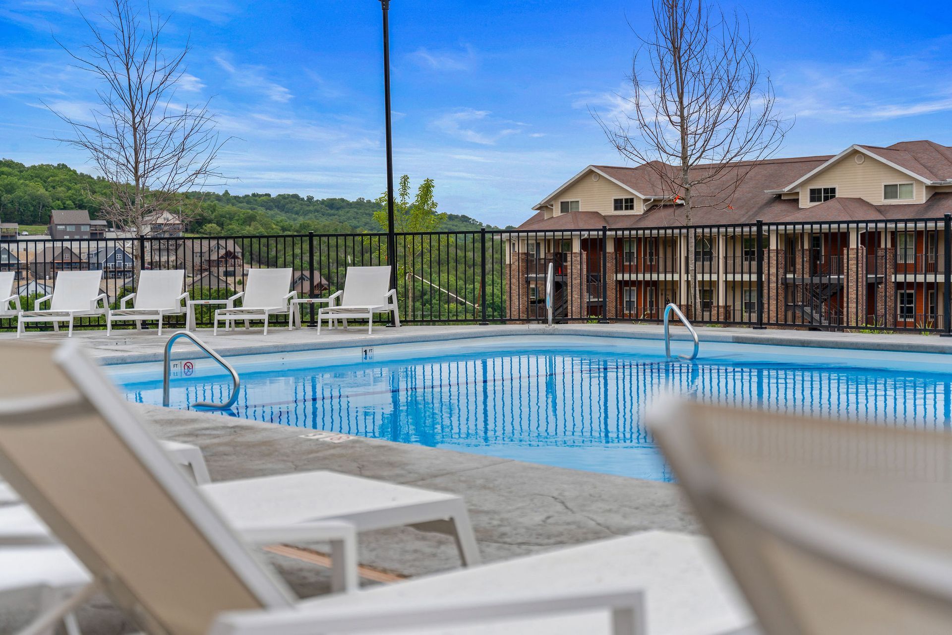 Outdoor apartment pool with lounge chairs and a fence, viewed from a patio.