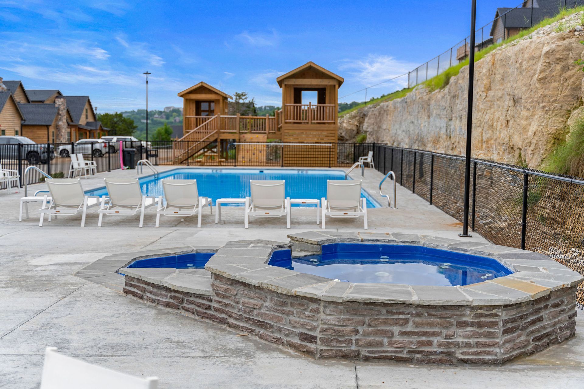 Poolside hot tub beside an outdoor swimming pool with lounge chairs and wooden cabanas in a resort setting
