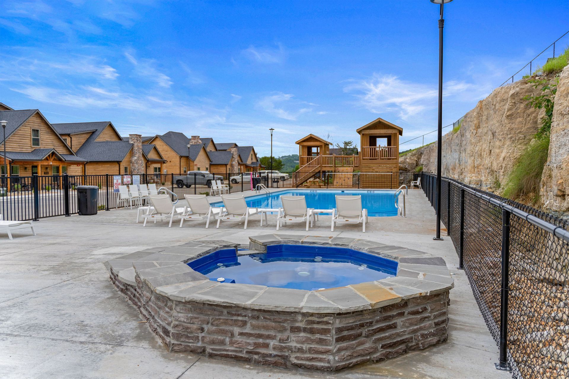 Outdoor pool area with a stone hot tub, lounge chairs, and cabins under a blue sky