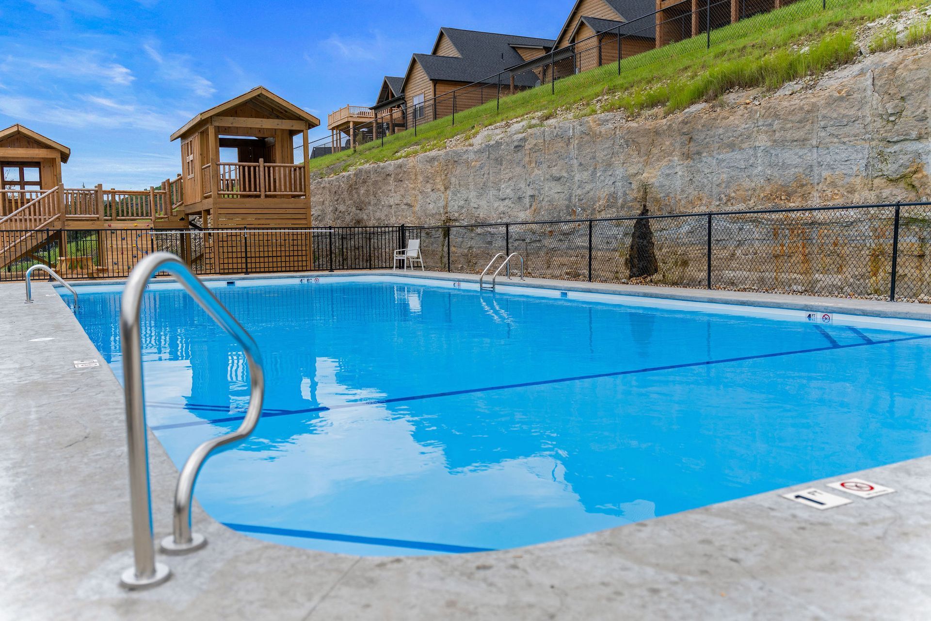 Empty blue swimming pool with metal handrail beside stone retaining wall and hillside cabins