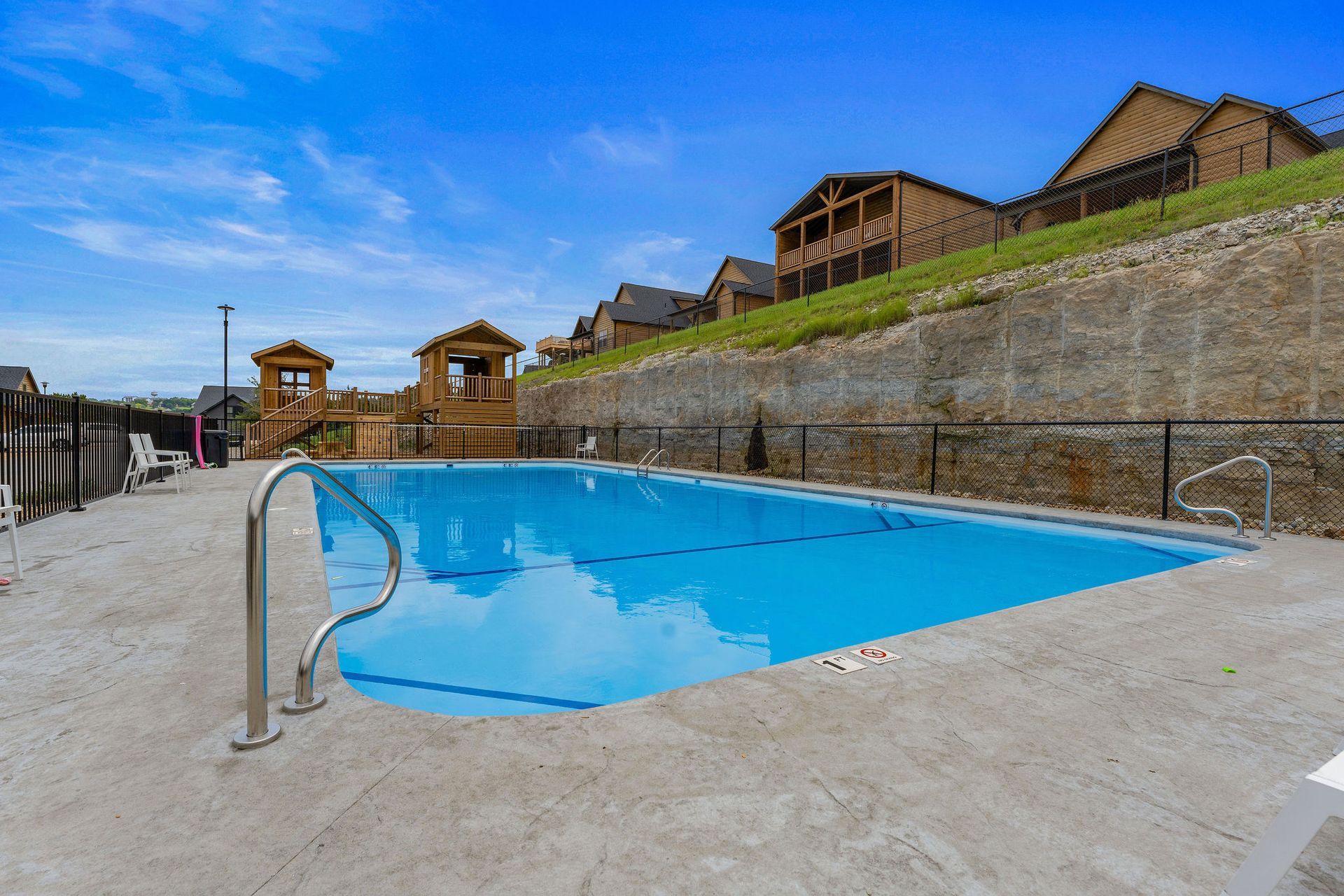 Empty outdoor swimming pool with blue water, concrete deck, and wooden cabins on a hillside behind it